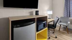 Hotelkamer met een flatscreen televisie aan de muur, een koffiezetapparaat op een houten plank, een bureau met een lamp, een zwarte bureaustoel en een bed met een grijs hoofdeinde.
