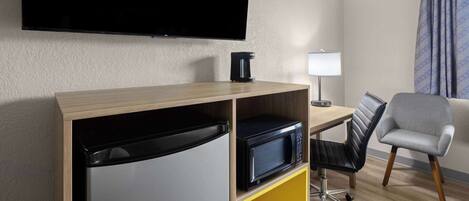 Habitación de hotel con televisor de pantalla plana en la pared, cafetera sobre una estantería de madera, escritorio con lámpara, silla de oficina negra y cama con cabecero gris.