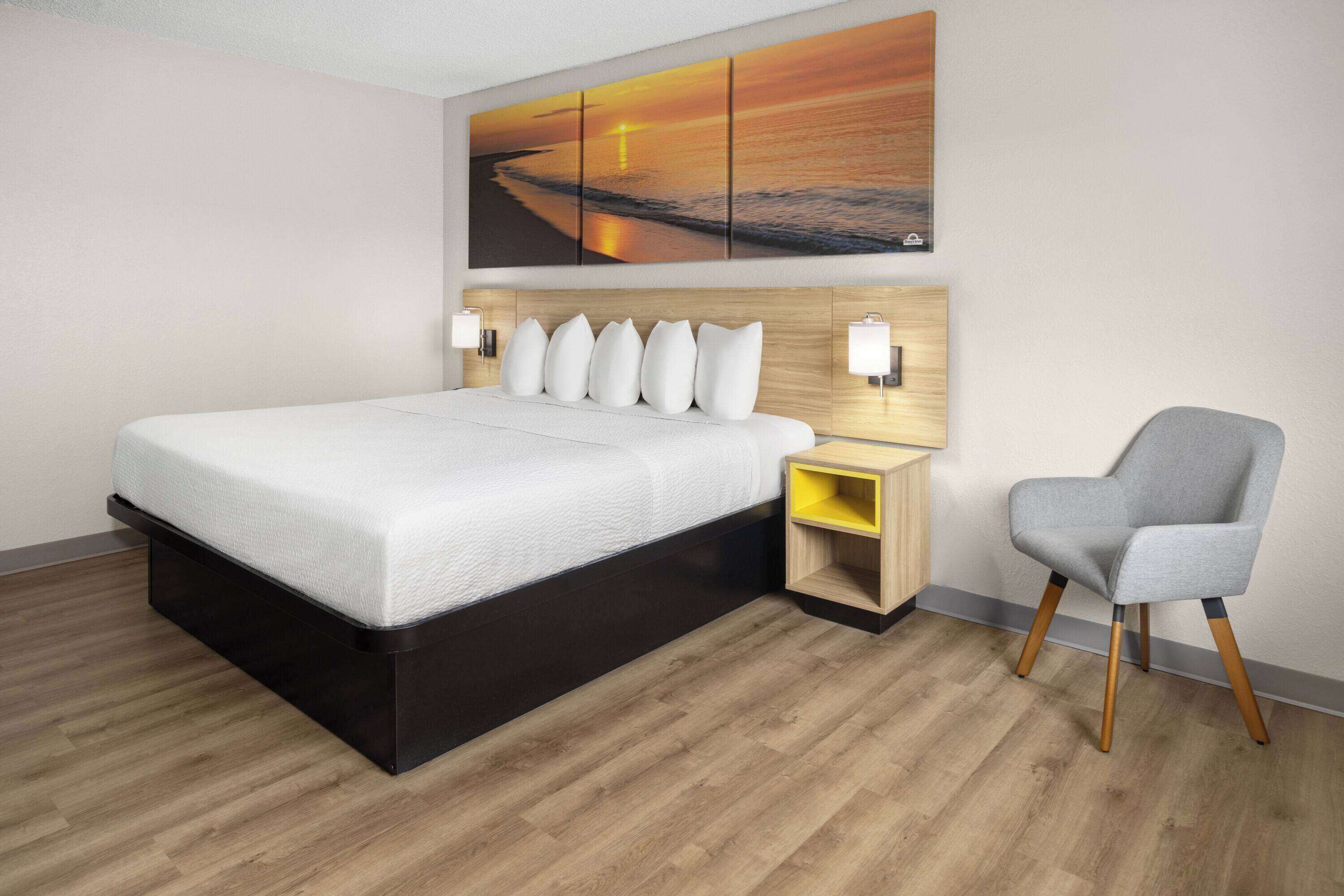 Quarto de hotel moderno com uma cama grande, uma mesa de cabeceira de madeira, uma cadeira e uma televisão fixada na parede.