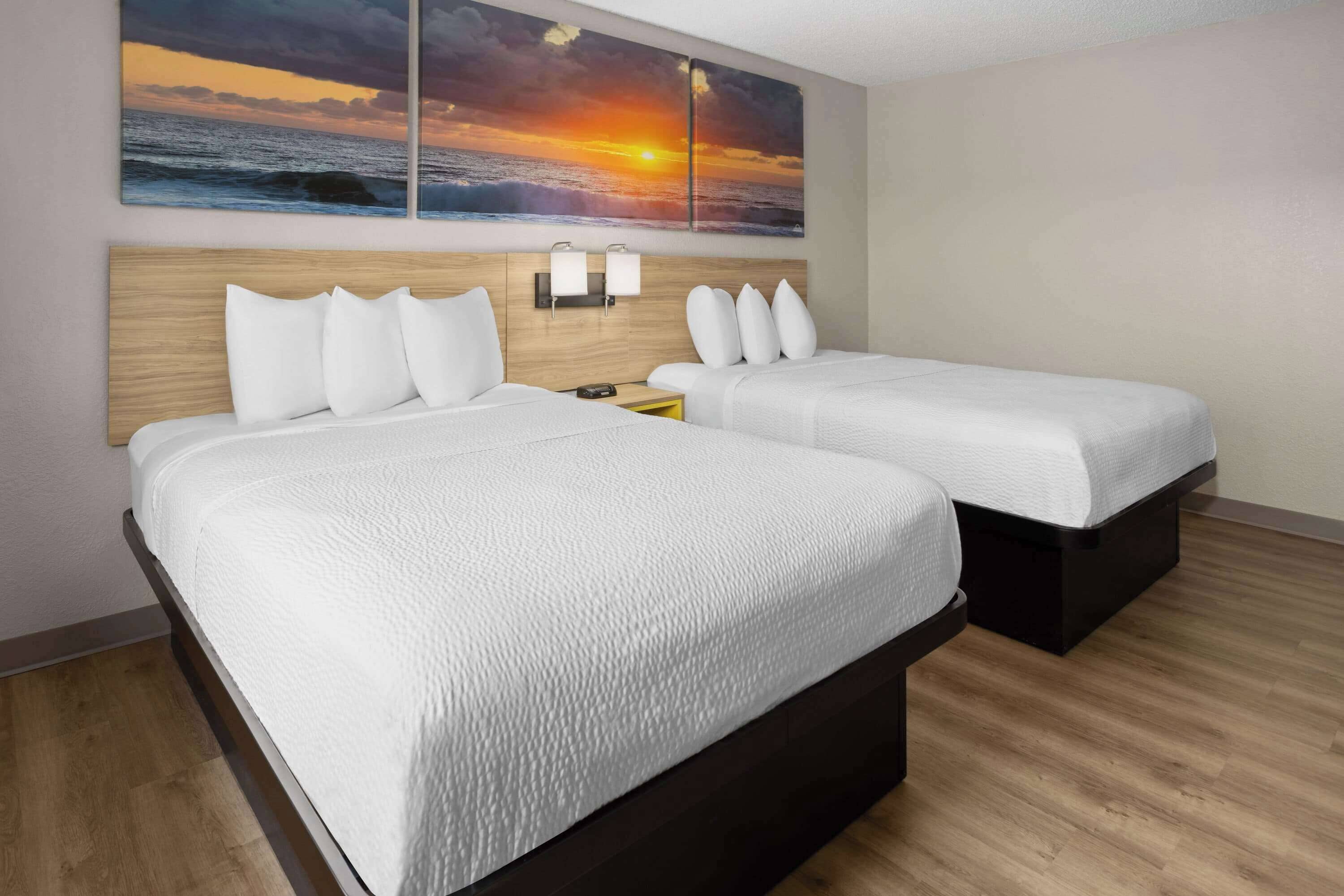 A hotel room with two beds, a wooden headboard, a wall-mounted light, and a sunset painting.