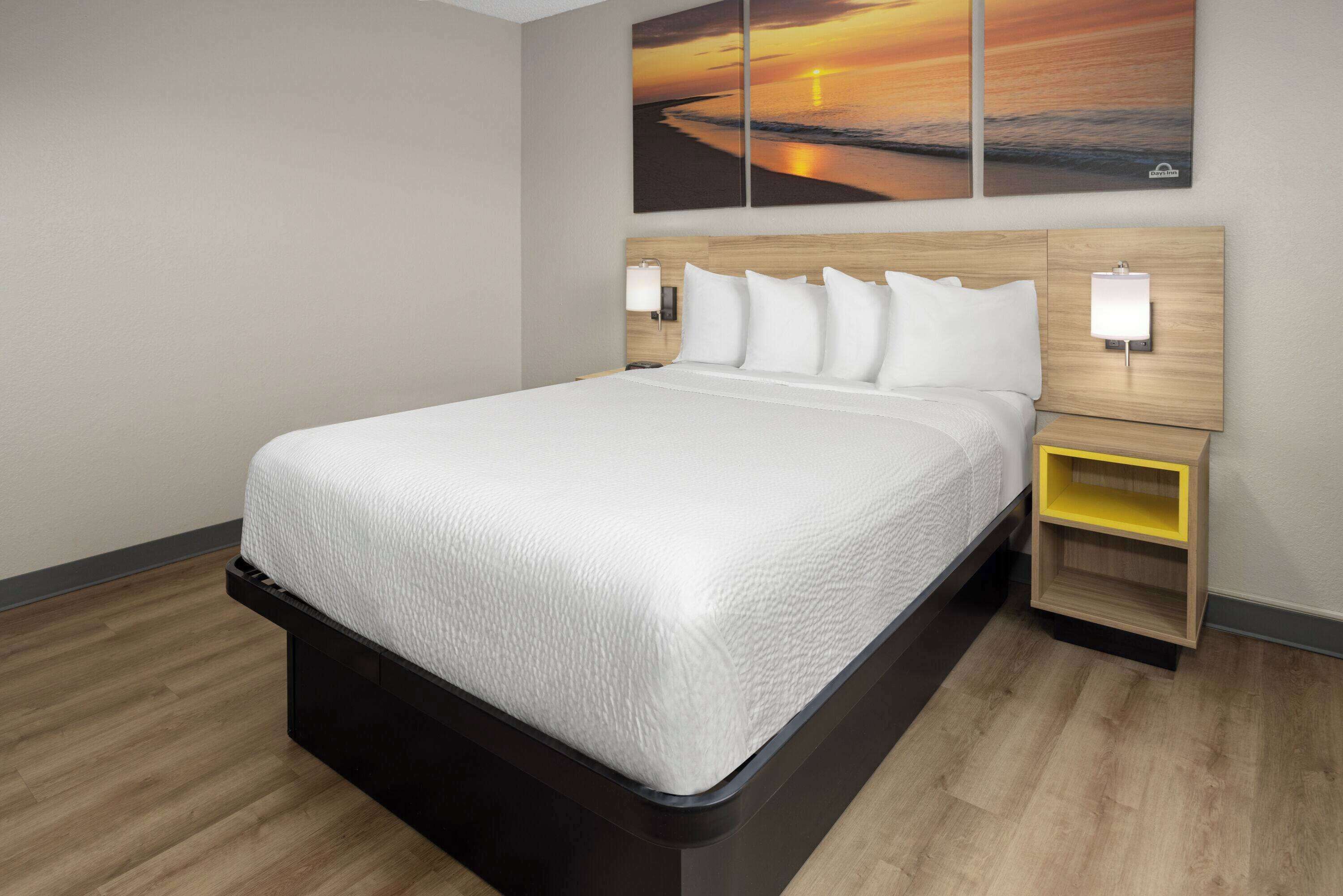 Ein Hotelzimmer mit einem großen Bett, einem Holz-Kopfteil, zwei Nachttischen mit Lampen und einem Wandbild, das einen Sonnenuntergang zeigt.
