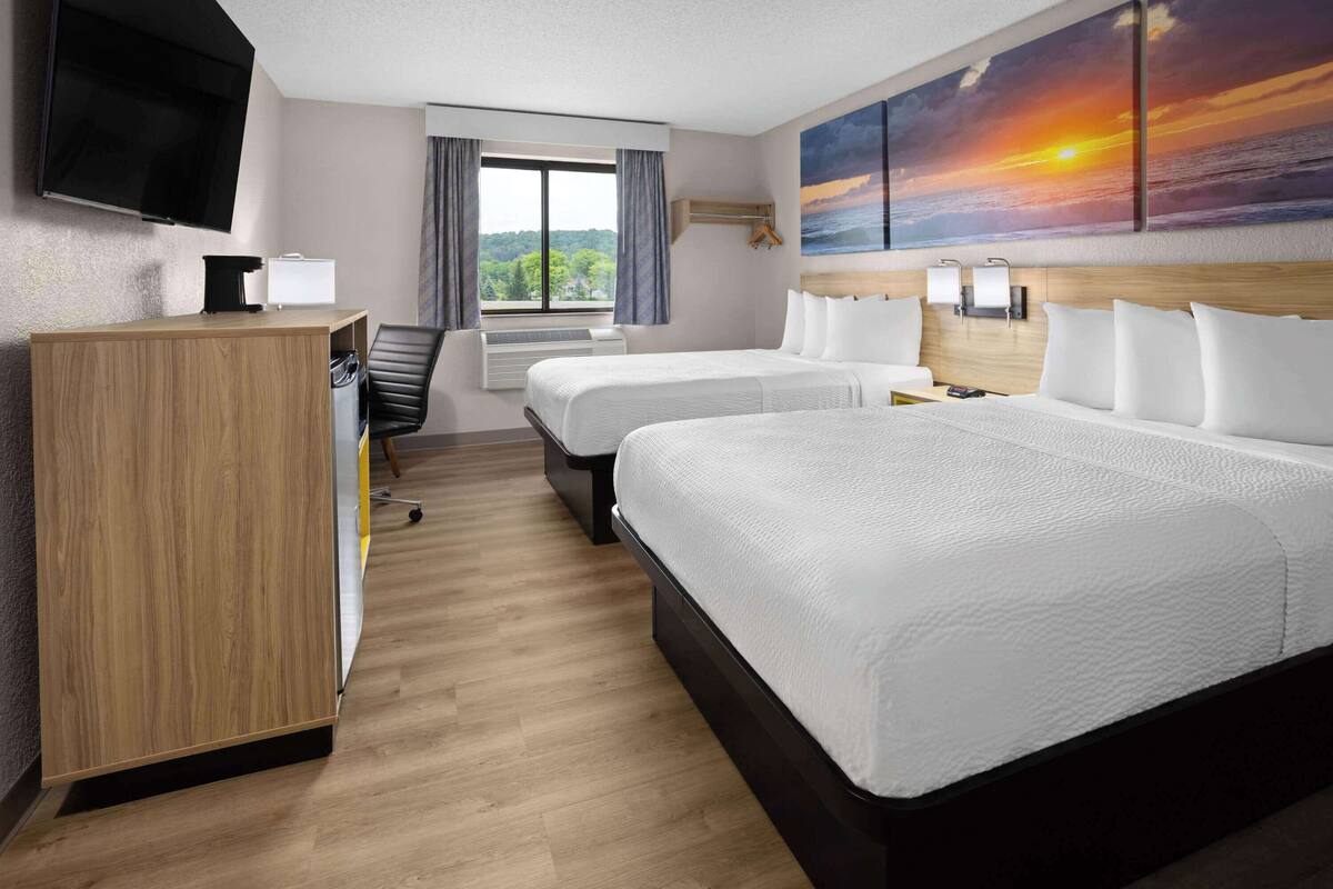 Une chambre d’hôtel avec deux lits, un bureau, une télévision, une fenêtre avec des rideaux et un tableau représentant un coucher de soleil accroché au mur.
