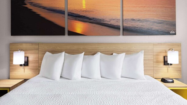 Een hotelkamer met een groot schilderij van een zonsondergang, een houten hoofdbord, wit beddengoed en nachtlampjes.