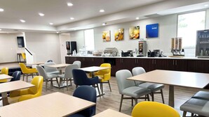 A modern dining area with multiple tables and chairs, a kitchen counter with coffee machines, and artwork on the walls.