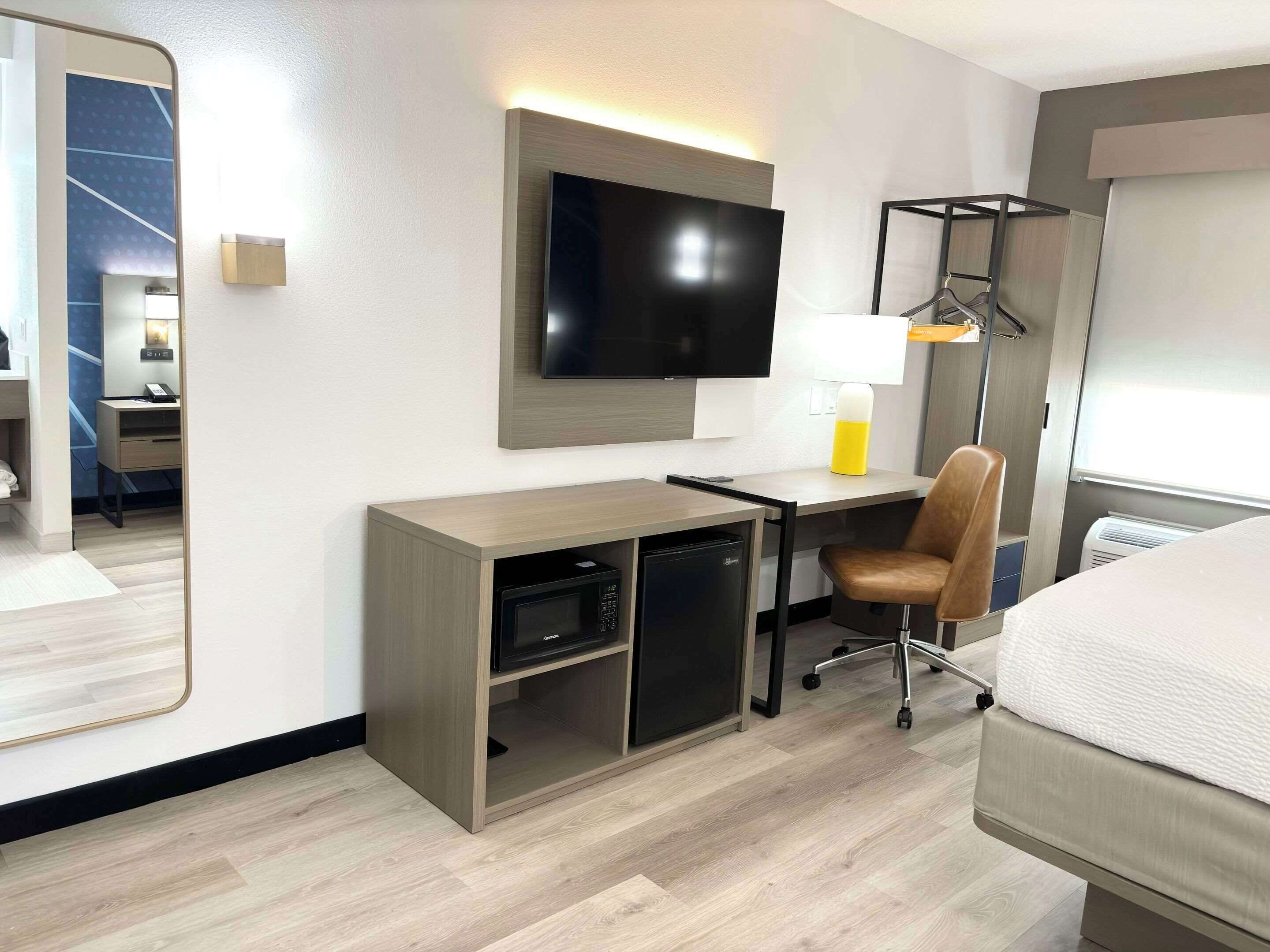A hotel room with a bed, a desk with a computer, a chair, a TV mounted on the wall, and a lamp.
