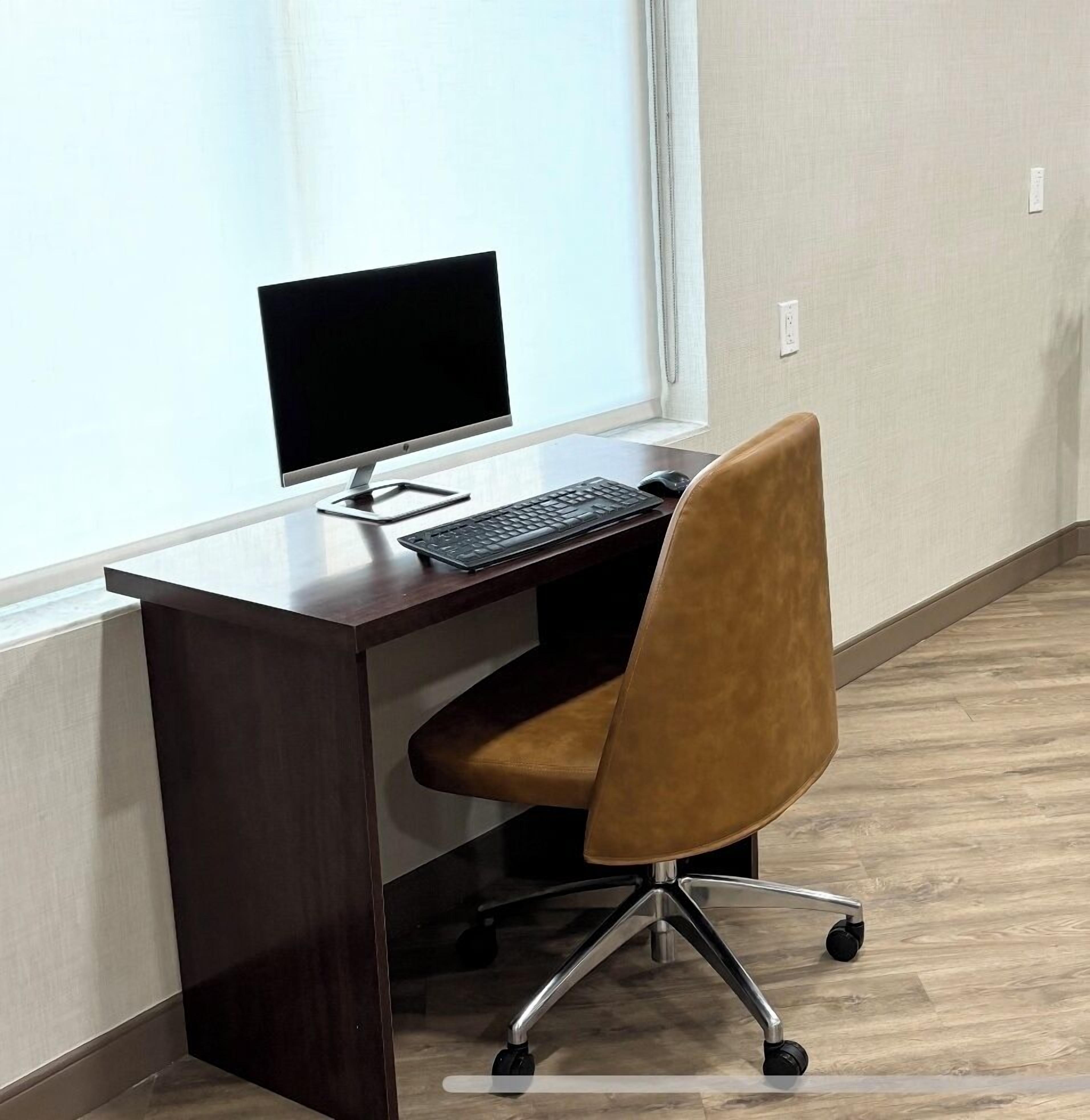 Un escritorio de madera con una computadora y teclado, una silla de oficina marrón y una ventana con cortinas.