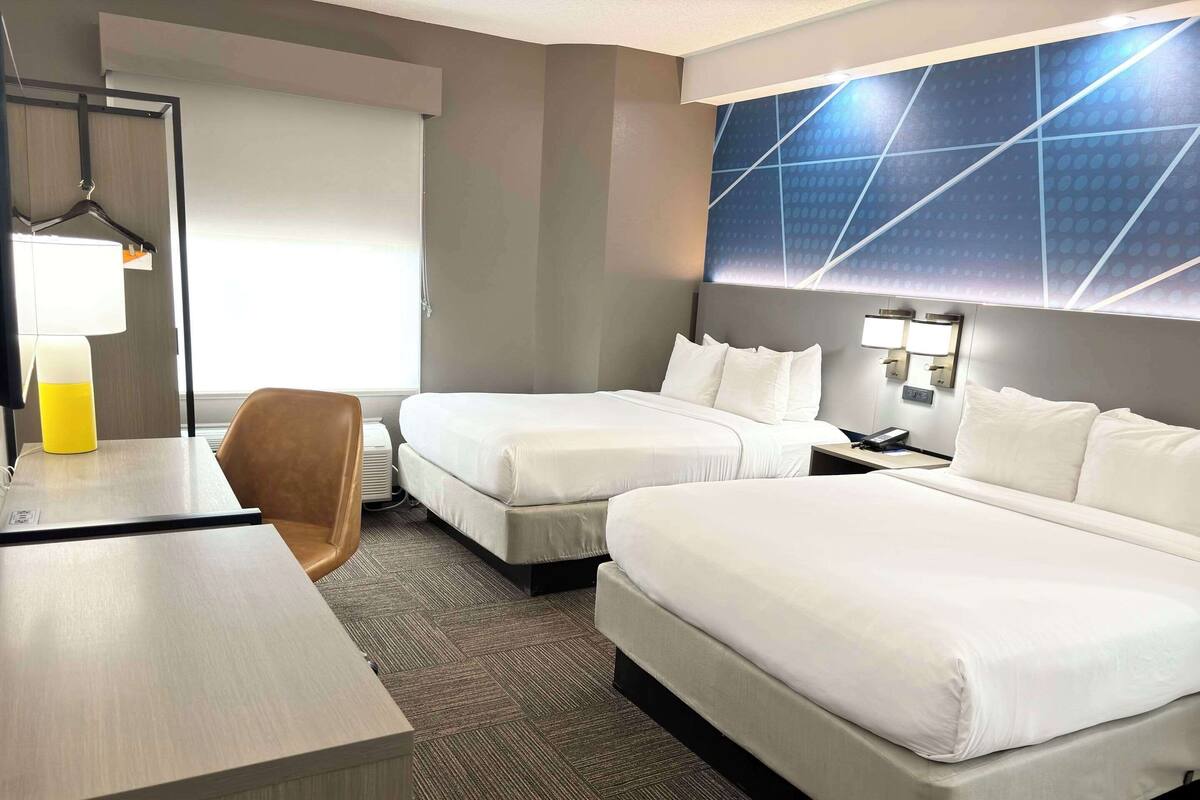 A hotel room with two beds, a desk, a chair, and a large wall with a geometric pattern.