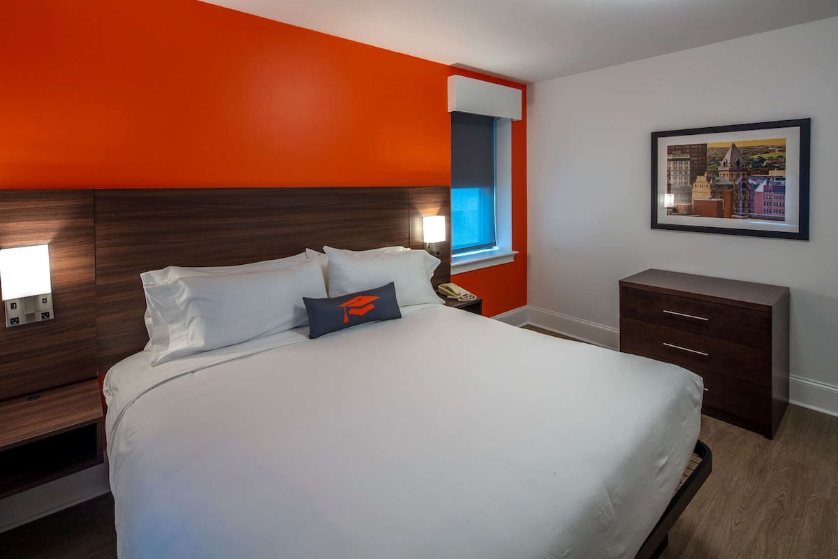 Habitación de hotel con una cama grande, una mesita de noche con lámpara, un aparador y un cuadro con una vista urbana enmarcada en la pared.