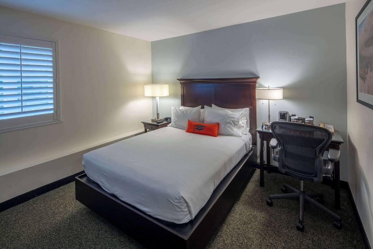 Una habitación de hotel con una cama grande, dos lámparas de noche, un escritorio con una silla y una ventana con persianas.