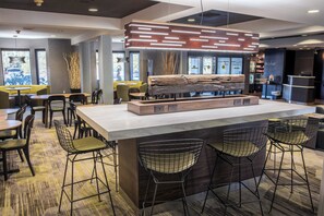 A modern bar area with a central island, bar stools, and a seating area with tables and chairs. - Courtyard by Marriott Norwich (Norwich)
