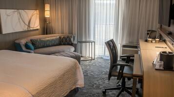 Ein Hotelzimmer mit einem großen Bett, einem Schreibtisch, einem Sessel, einem Fernseher und einem Fenster mit Vorhängen.