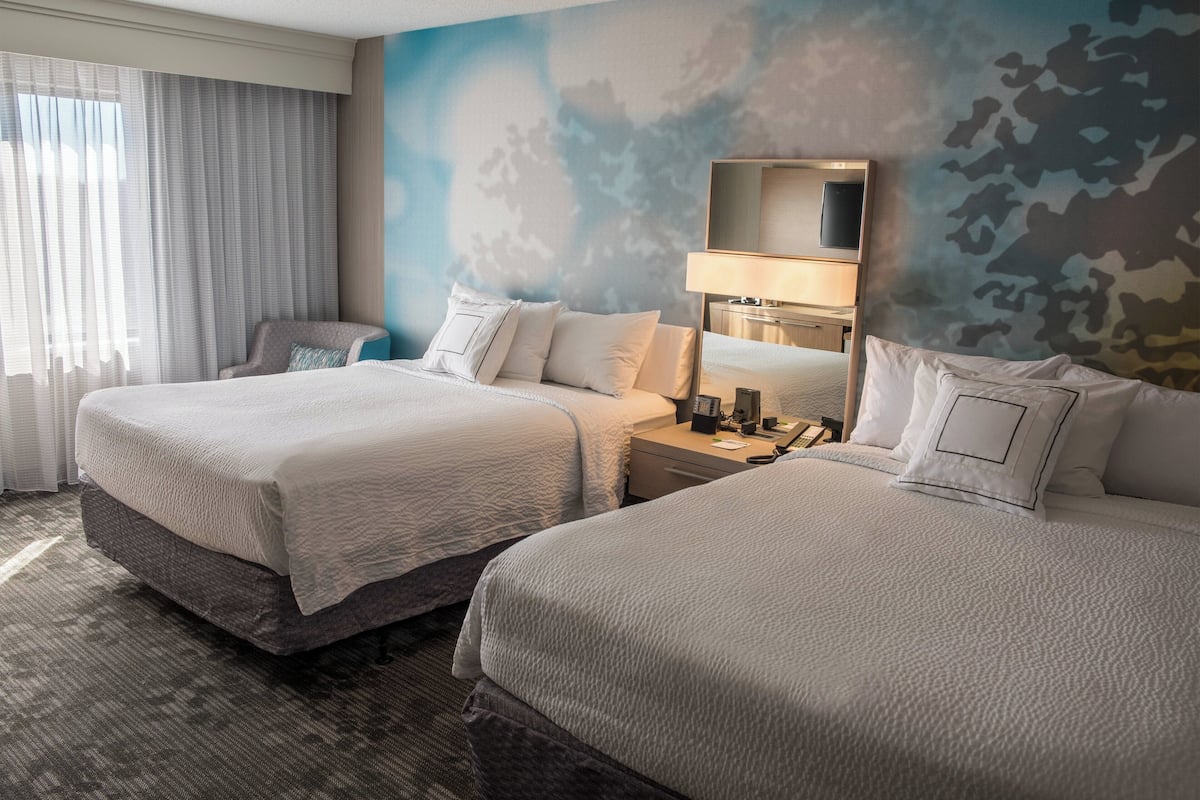 A hotel room with two beds, a chair, a nightstand with a lamp, and a large window with curtains.