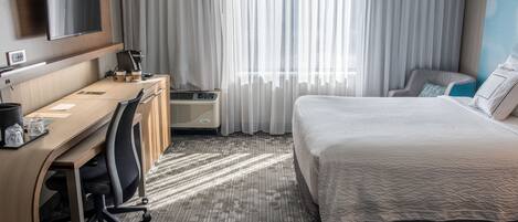 Habitación de hotel con una cama, un escritorio con silla, televisión en la pared y una ventana con cortinas.