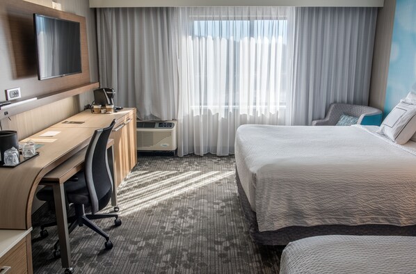A hotel room with a bed, a desk with a chair, a television mounted on the wall, and a window with curtains.