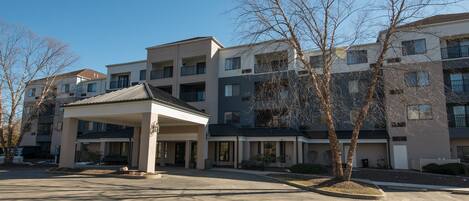 A multi-story residential building with a covered entrance, multiple balconies, and a parking area in front.