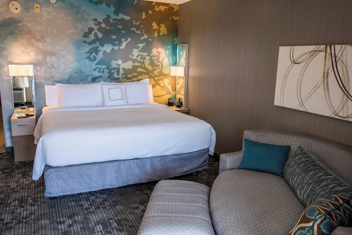 A hotel room with a large bed, a sofa, a nightstand with a lamp, and a wall with abstract art.