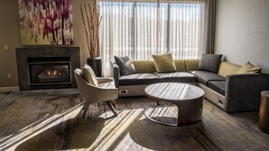 Ein modernes Wohnzimmer mit Kamin, Ecksofa, rundem Couchtisch und einem großen Fenster mit Vorhängen.