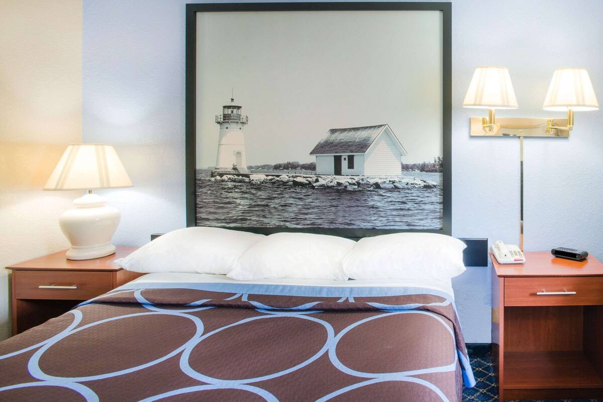 Ein Hotelzimmer mit einem Bett, zwei Nachttischen, einer Lampe, einem Gemälde von einem Leuchtturm und einem Haus sowie einem Telefon.