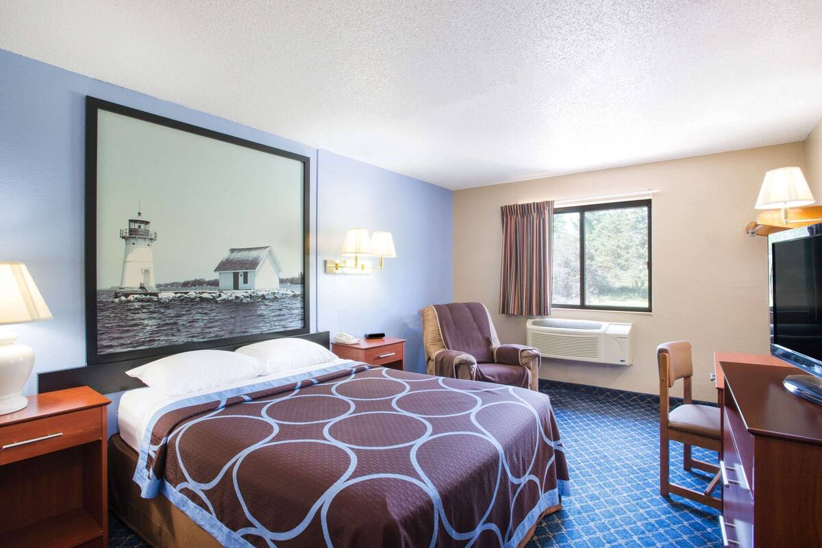 Ein Hotelzimmer mit einem großen Bett, einem Schreibtisch, einem Stuhl, einem Fernseher, einem Fenster mit Vorhängen und einem Gemälde eines Leuchtturms.