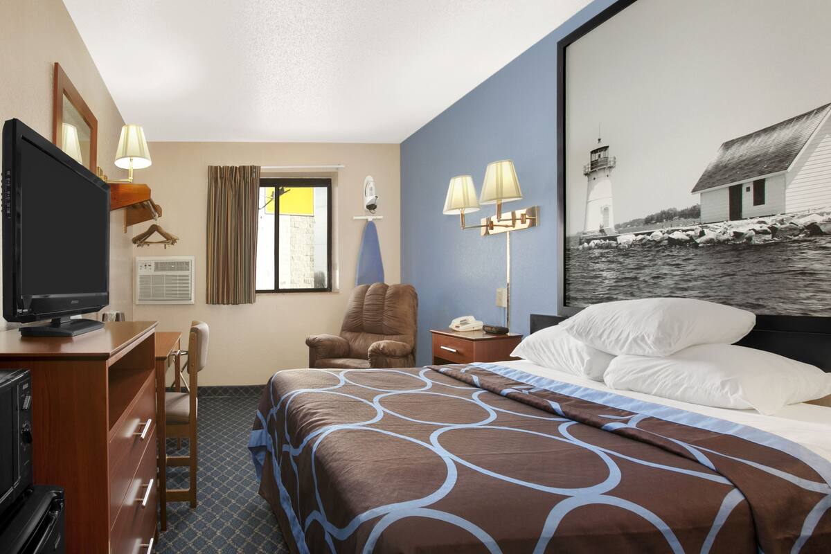 Ein Hotelzimmer mit einem Bett, einem Schreibtisch mit Fernseher, einem Sessel, einem Nachttisch mit Lampen und einem Gemälde eines Leuchtturms.