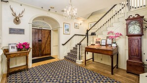 Un vaste hall d’entrée agrémenté d’un tapis à motifs, d’un escalier en bois, d’un lustre, d’une tête de cerf fixée au mur et d’une grande horloge.