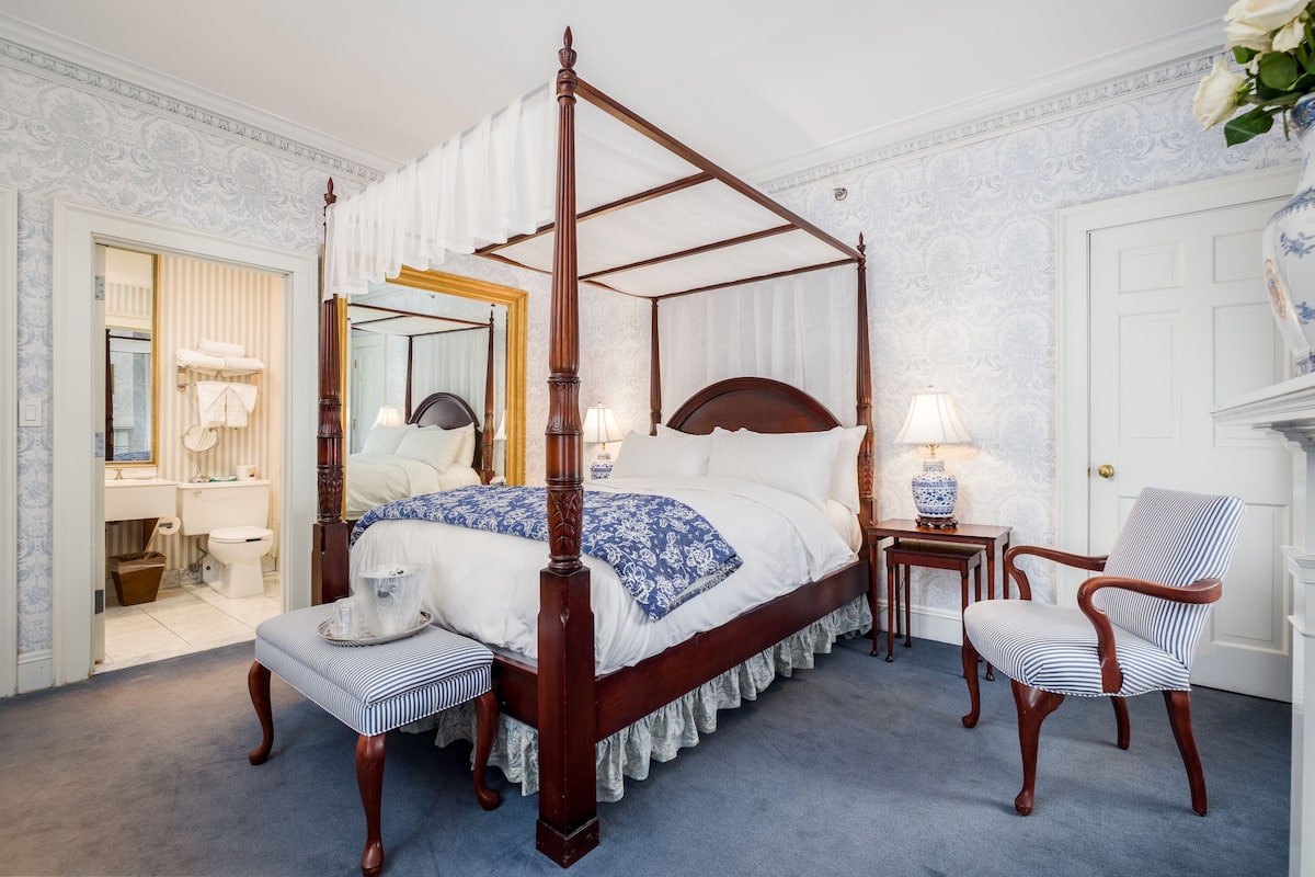 A four-poster bed with a canopy, a bedside table with a lamp, a chair, and a bench.