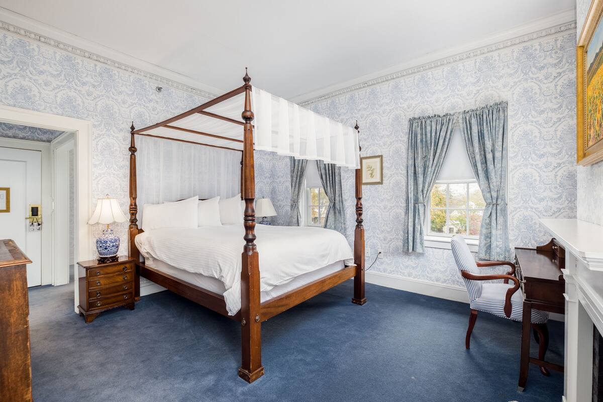 A four-poster bed with white linens, a wooden nightstand with a lamp, a striped chair, and a desk with a mirror.