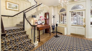 Un magnifique escalier recouvert d’une moquette à motifs, une table en bois ornée d’un vase de fleurs, un lustre et une horloge.