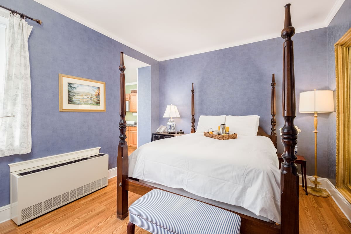 A four-poster bed in a room with a blue wall, a radiator, a bedside table with a lamp, and a window with curtains.