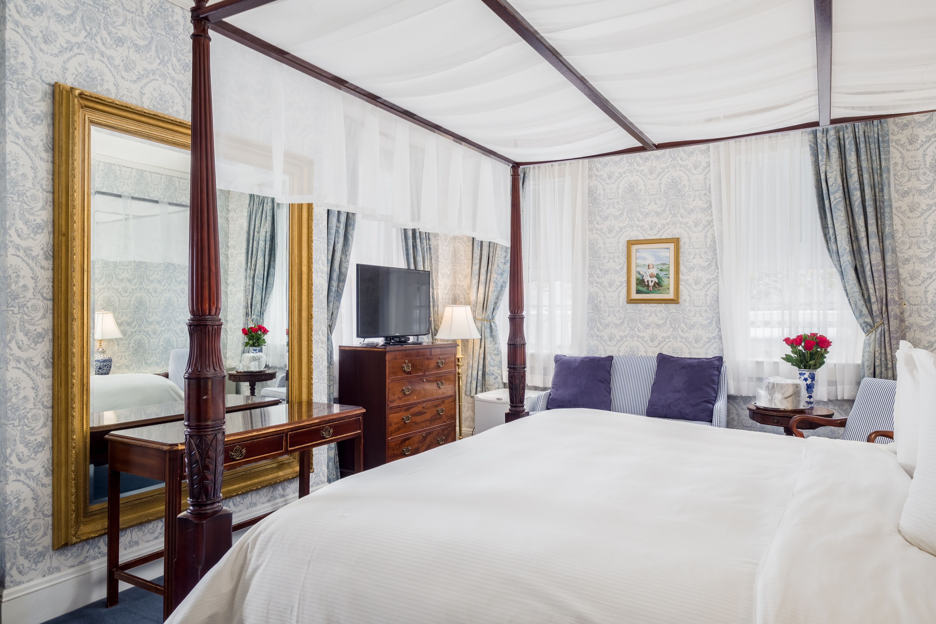 A four-poster bed with a large mirror, a television on a wooden dresser, and a window with curtains.