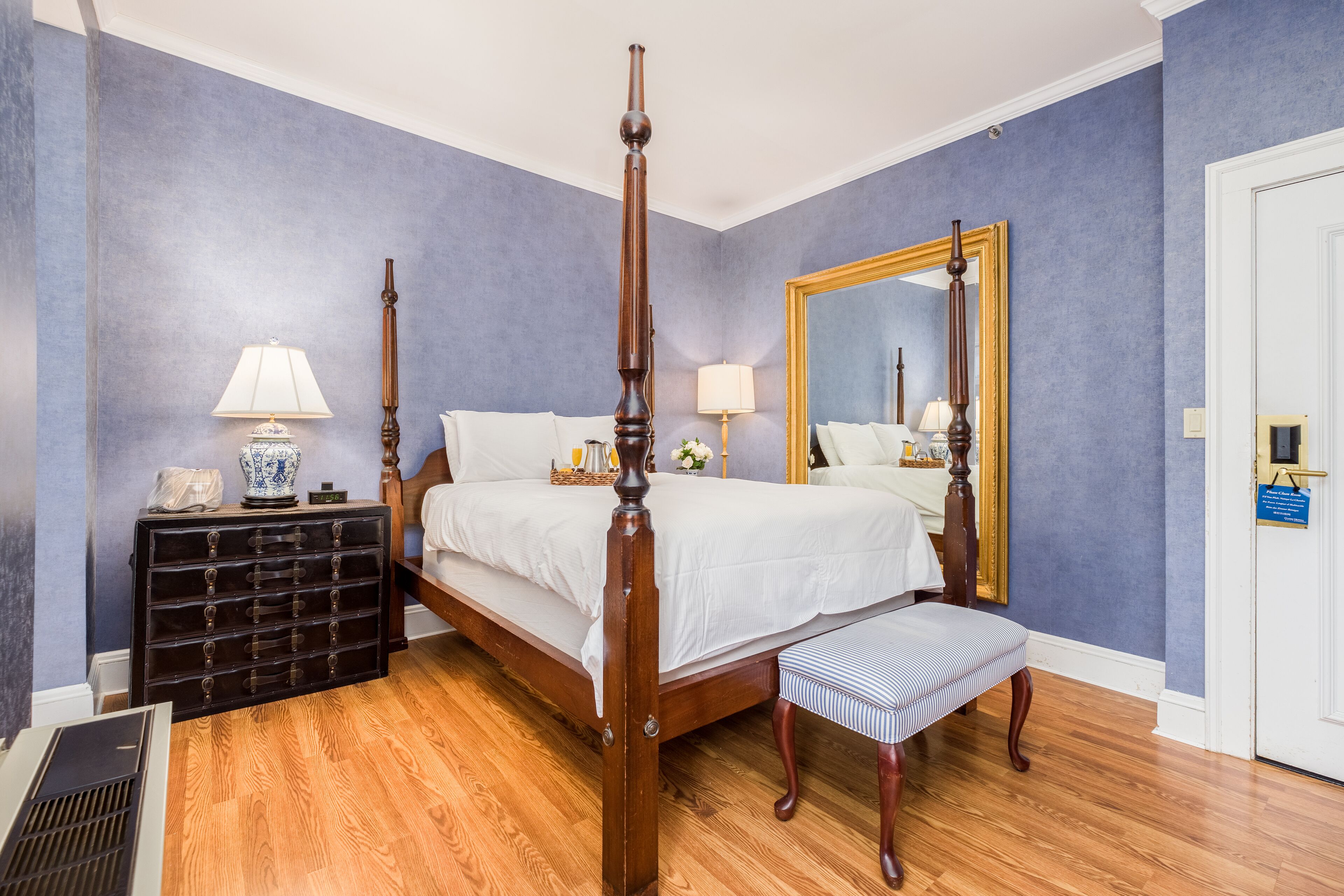 Una cama con dosel de madera, un banco al pie de la cama, un aparador con cajones y un espejo en la pared.