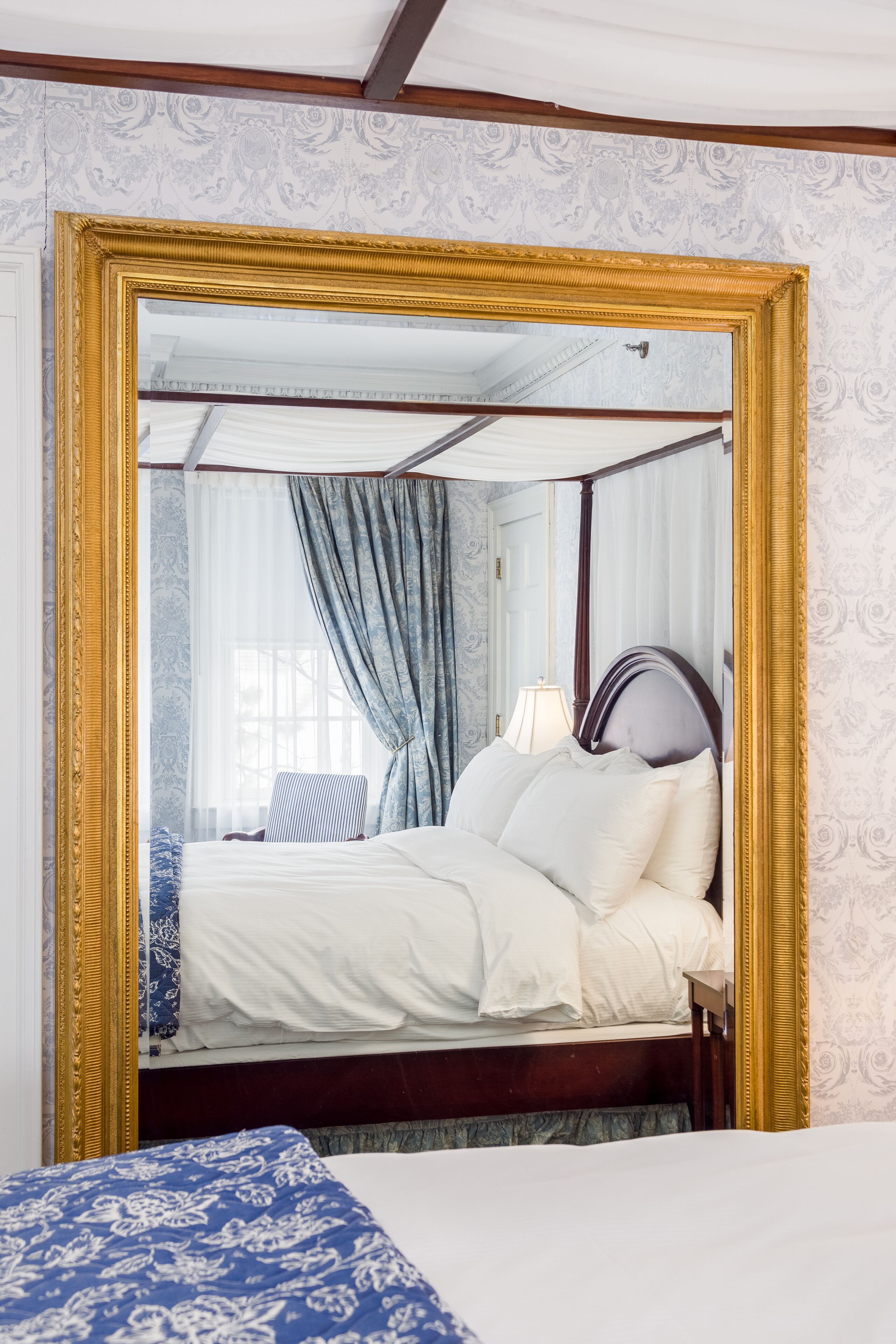 A bedroom with a large mirror, a bed with white linens, a blue patterned bedspread, and a wooden headboard.