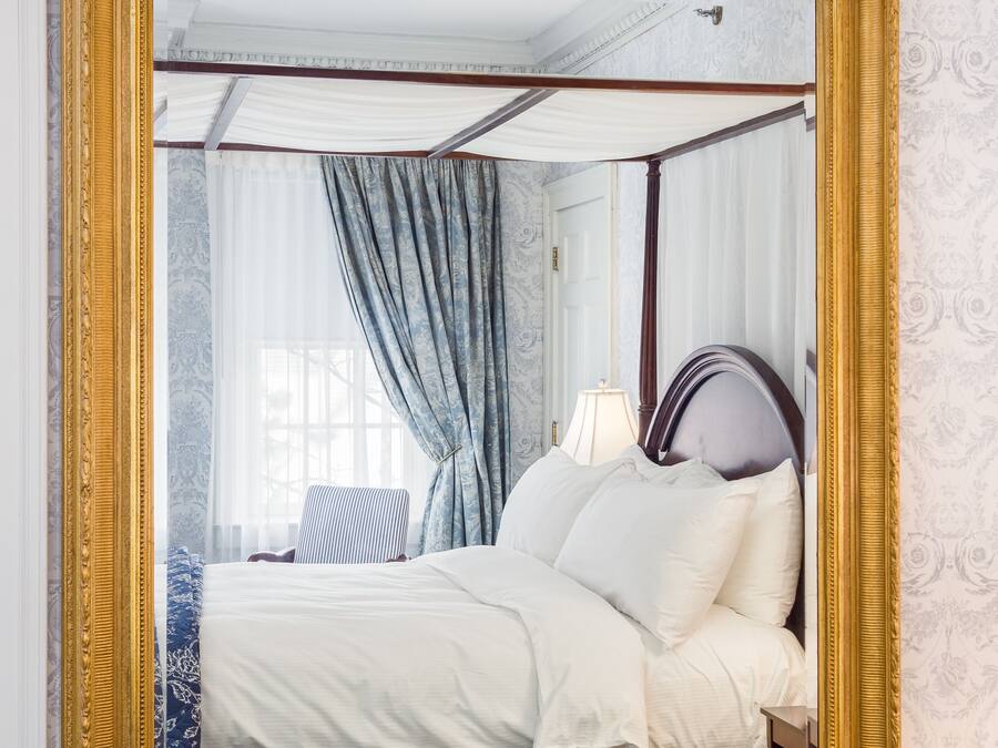 A bedroom with a large mirror, a bed with white linens, a blue patterned bedspread, and a wooden headboard.