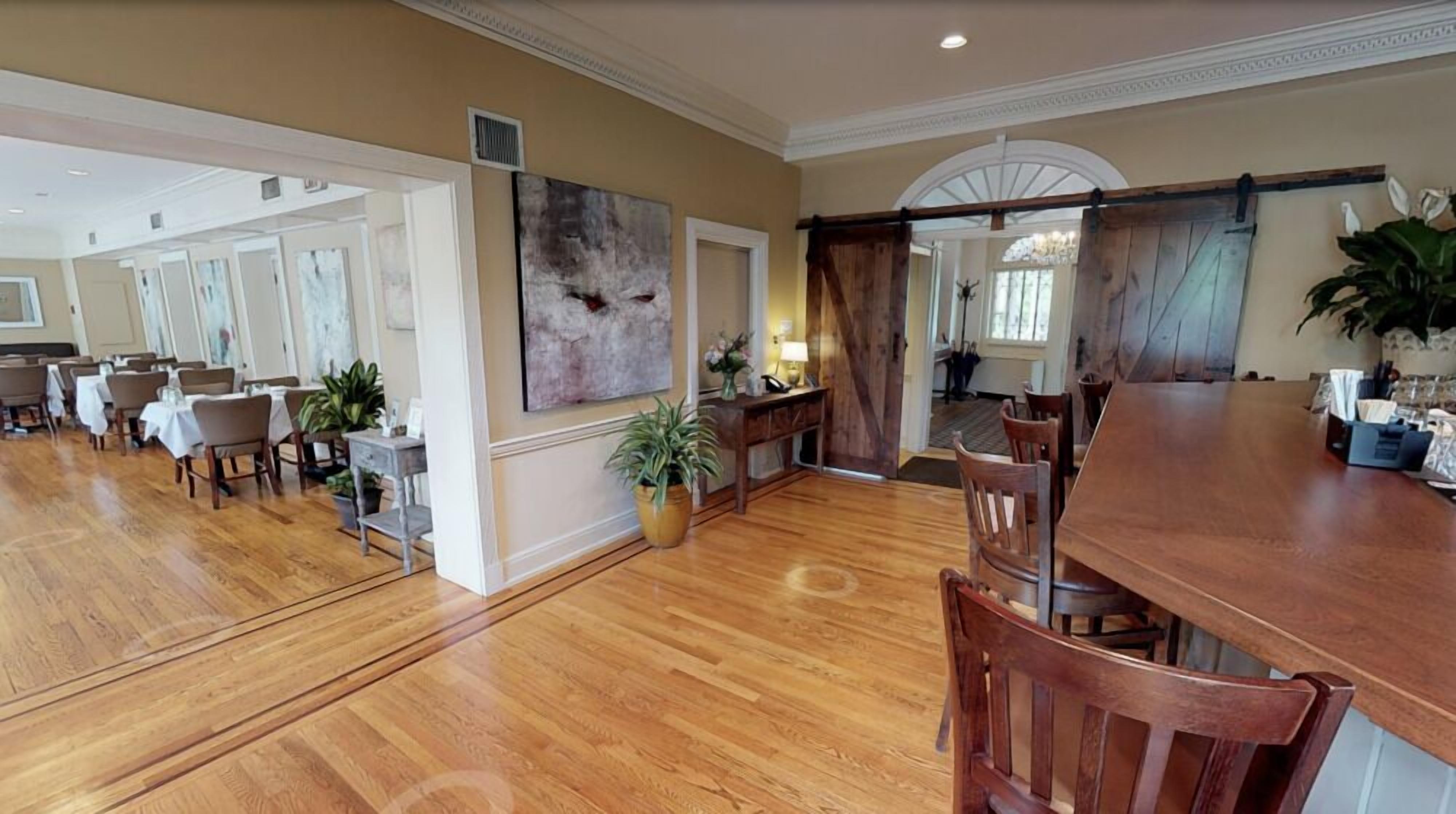 A spacious dining area with wooden flooring, a long table, and several chairs. There is a large painting on the wall and a potted plant on the floor.
