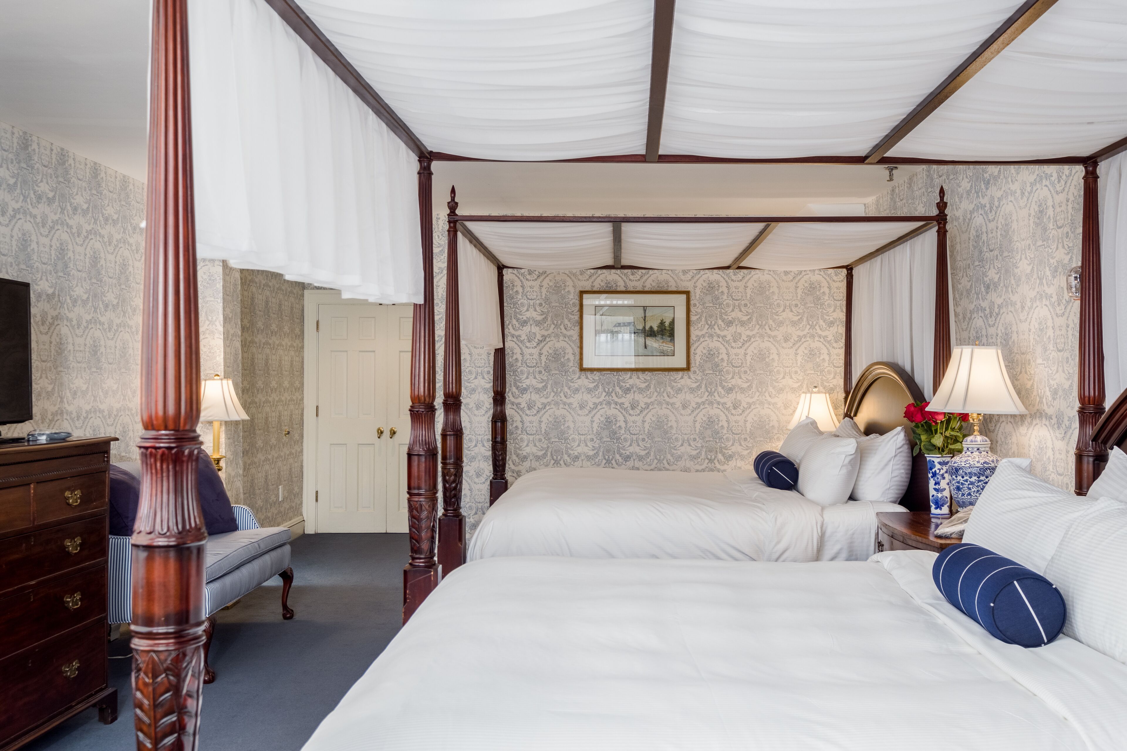 A four-poster bed with white linens, a wooden dresser, and a nightstand with a lamp.