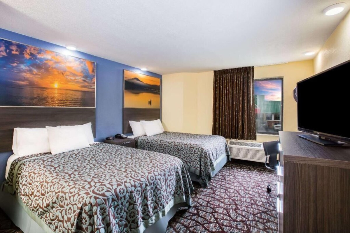 A hotel room with two beds, a flat-screen TV, a large window with curtains, and a painting of a sunset.