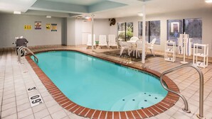 A pool area with a clear water surface, a lifeguard chair, and a wheelchair. There are white chairs and tables around the pool, and a window with a view of trees. - Days Inn by Wyndham Blue Springs (Blue Springs)