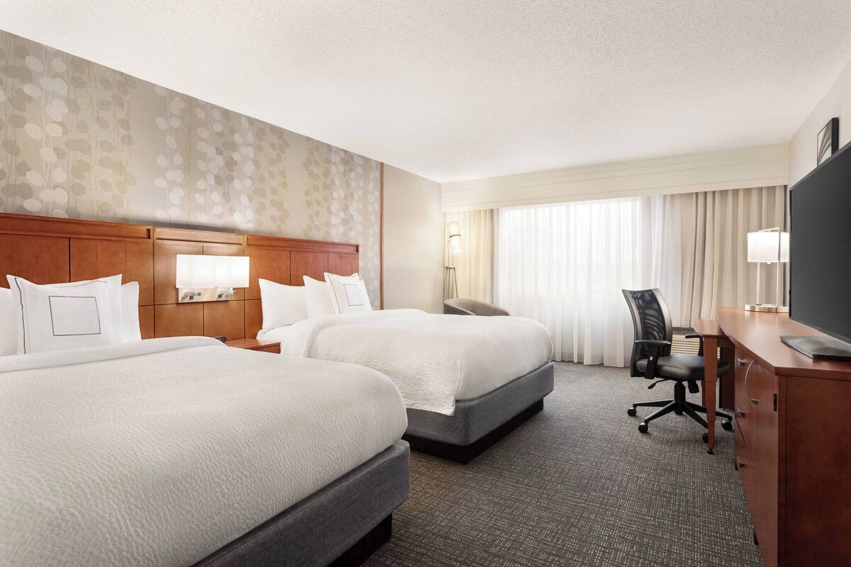 A hotel room with two beds, a desk, a chair, and a TV.