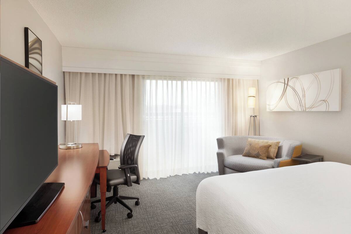 A hotel room with a bed, a desk with a laptop, a chair, a TV, a lamp, and a window with curtains.
