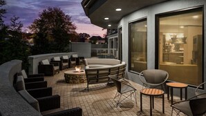 Un espace extérieur aménagé en terrasse, avec un foyer, des sièges et une vue sur l’intérieur grâce à de grandes fenêtres.