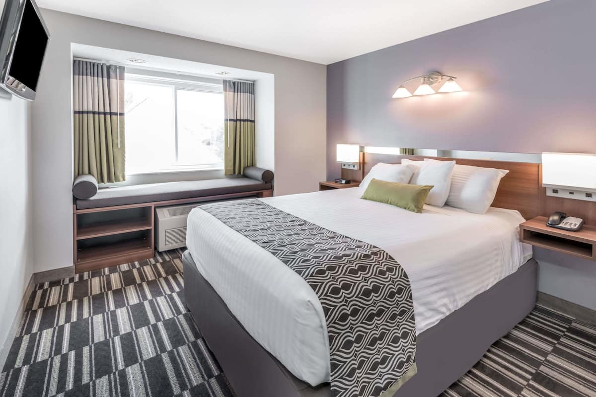 A hotel room with a large bed, bedside tables, a window with curtains, a TV, and a wall-mounted light fixture.