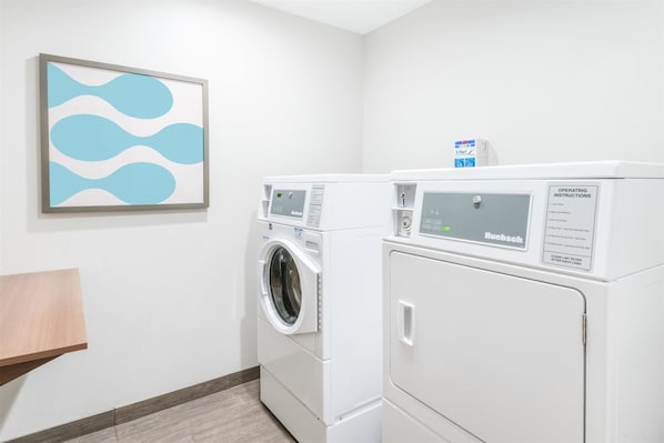 A laundry room with two white Huebsch washing machines, a wooden bench, and a framed abstract art piece on the wall.