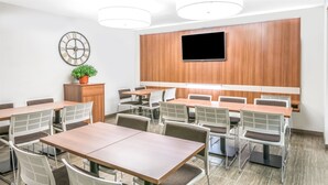 A conference room with multiple tables and chairs, a clock on the wall, a television mounted above the tables, and a potted plant on a cabinet. - Microtel Inn by Wyndham Victor/Rochester (Victor)