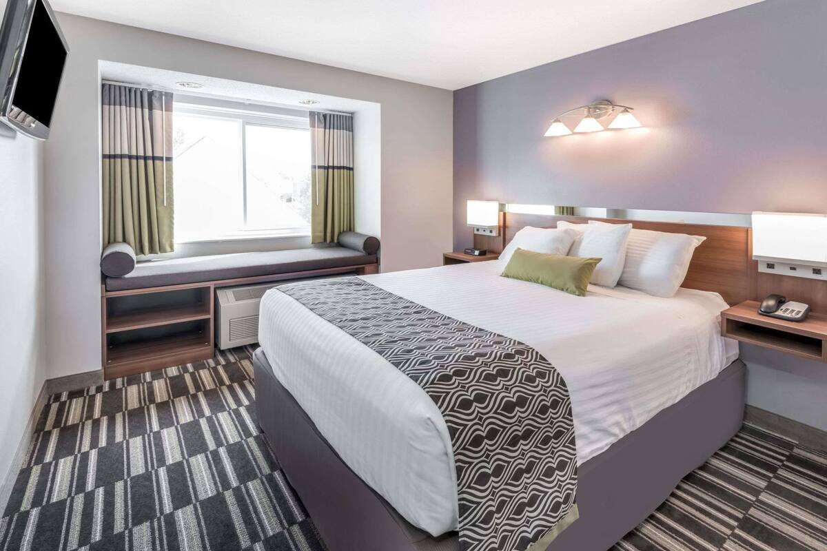 A hotel room with a large bed, a window with curtains, a bedside table with a lamp, and a wall-mounted TV.