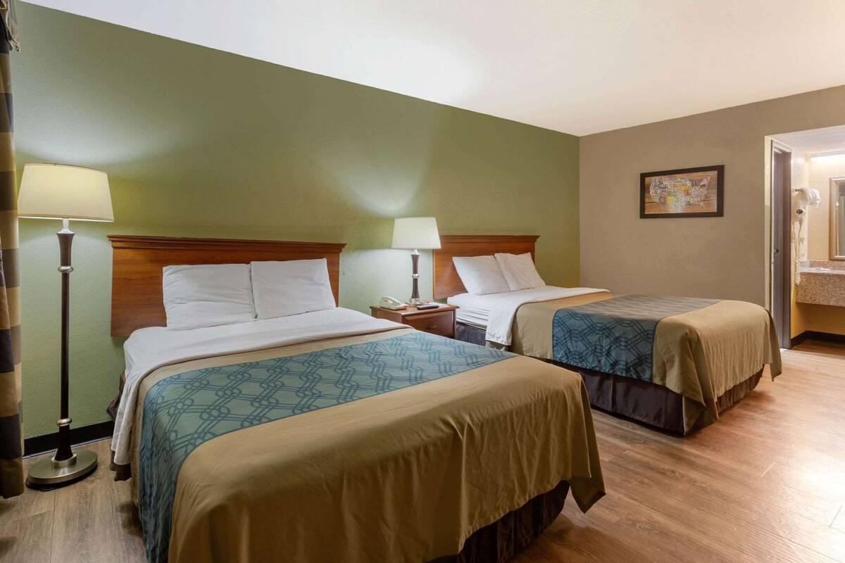 A hotel room with two beds, a nightstand with a lamp, a painting on the wall, and a bathroom visible through an open door.
