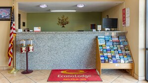 A reception desk with a red carpet, a flag, a wall-mounted flower, a computer, and a display of postcards. - Econo Lodge Belton - Kansas City South (Belton)