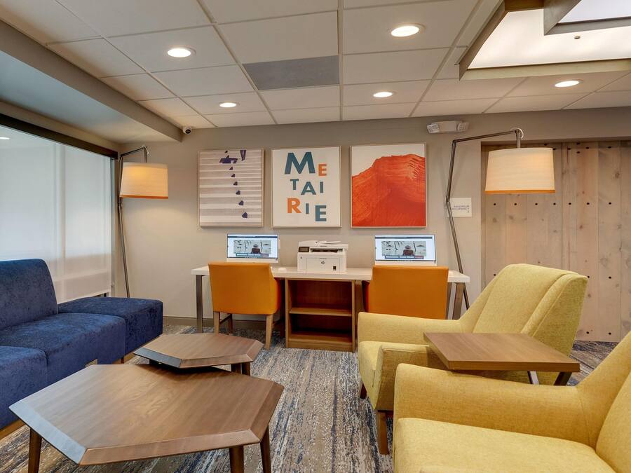 Una habitación de hotel moderna con un sofá azul, sillones amarillos, una mesa de centro de madera y un escritorio con dos computadoras portátiles y una impresora.