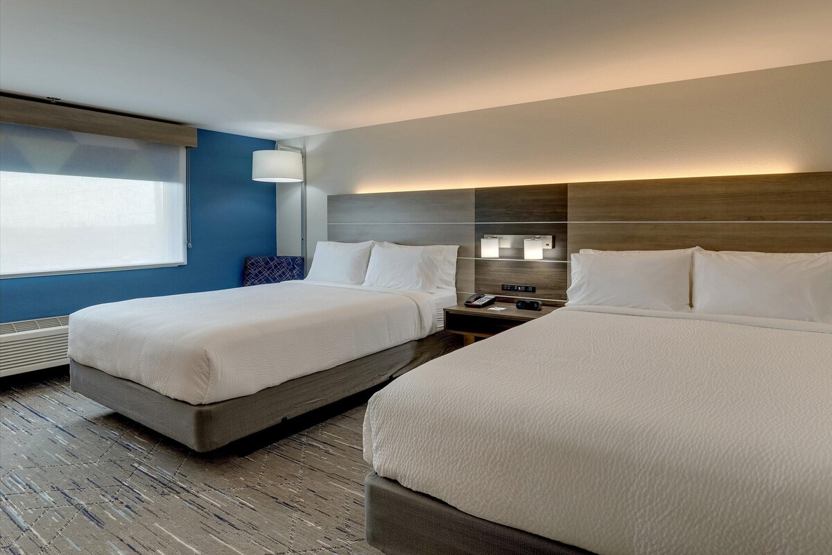 A hotel room with two beds, a bedside table with a phone and a lamp, and a window with blinds.