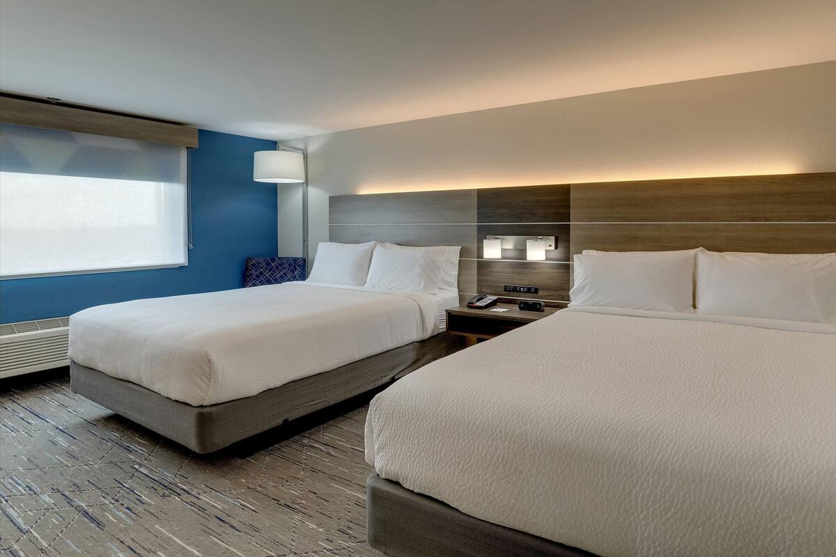 A hotel room with two beds, a bedside table with a phone and a lamp, and a window with blinds.