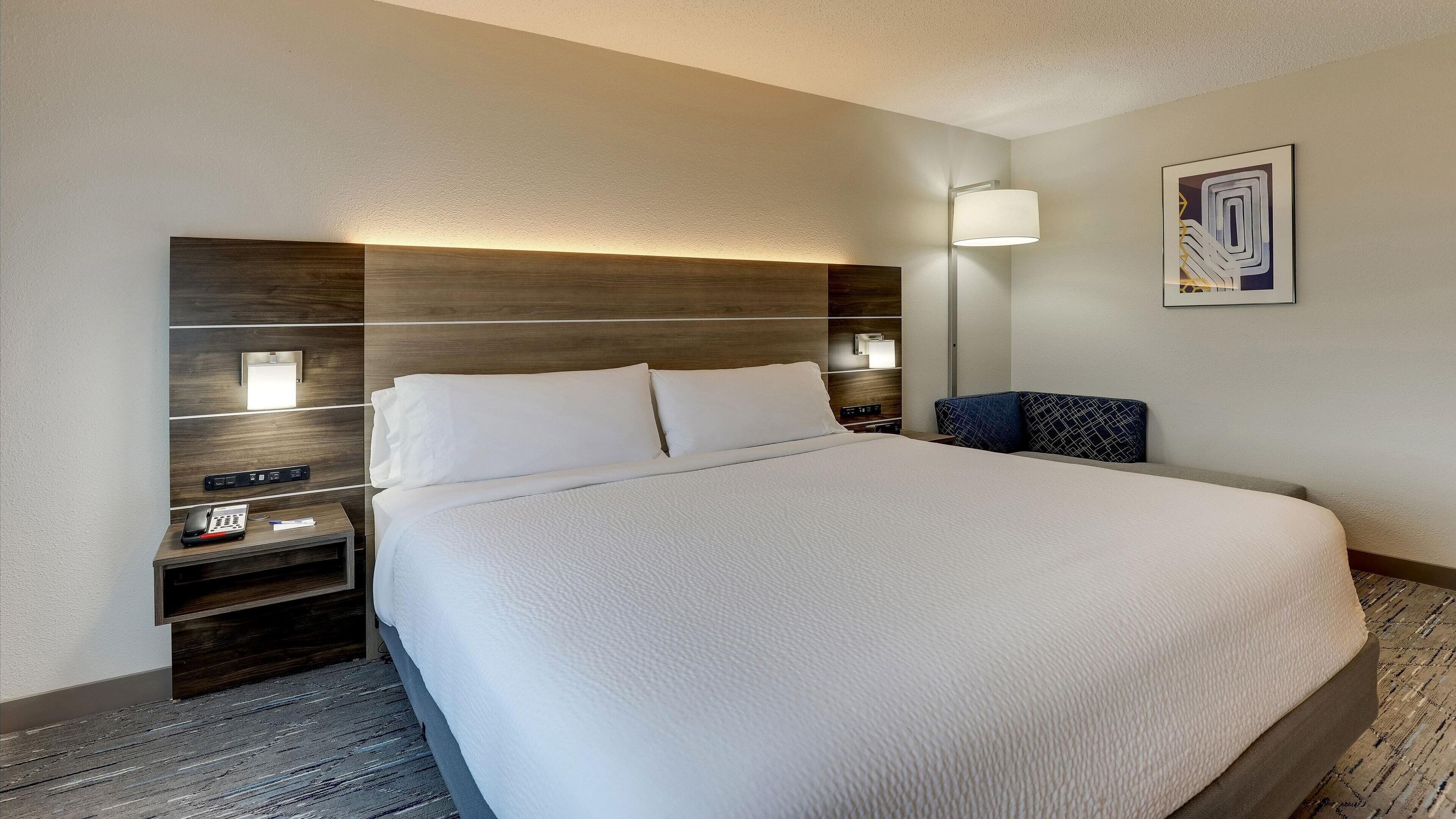 A hotel room with a large bed, a nightstand with a phone and a lamp, and a wall-mounted light fixture.